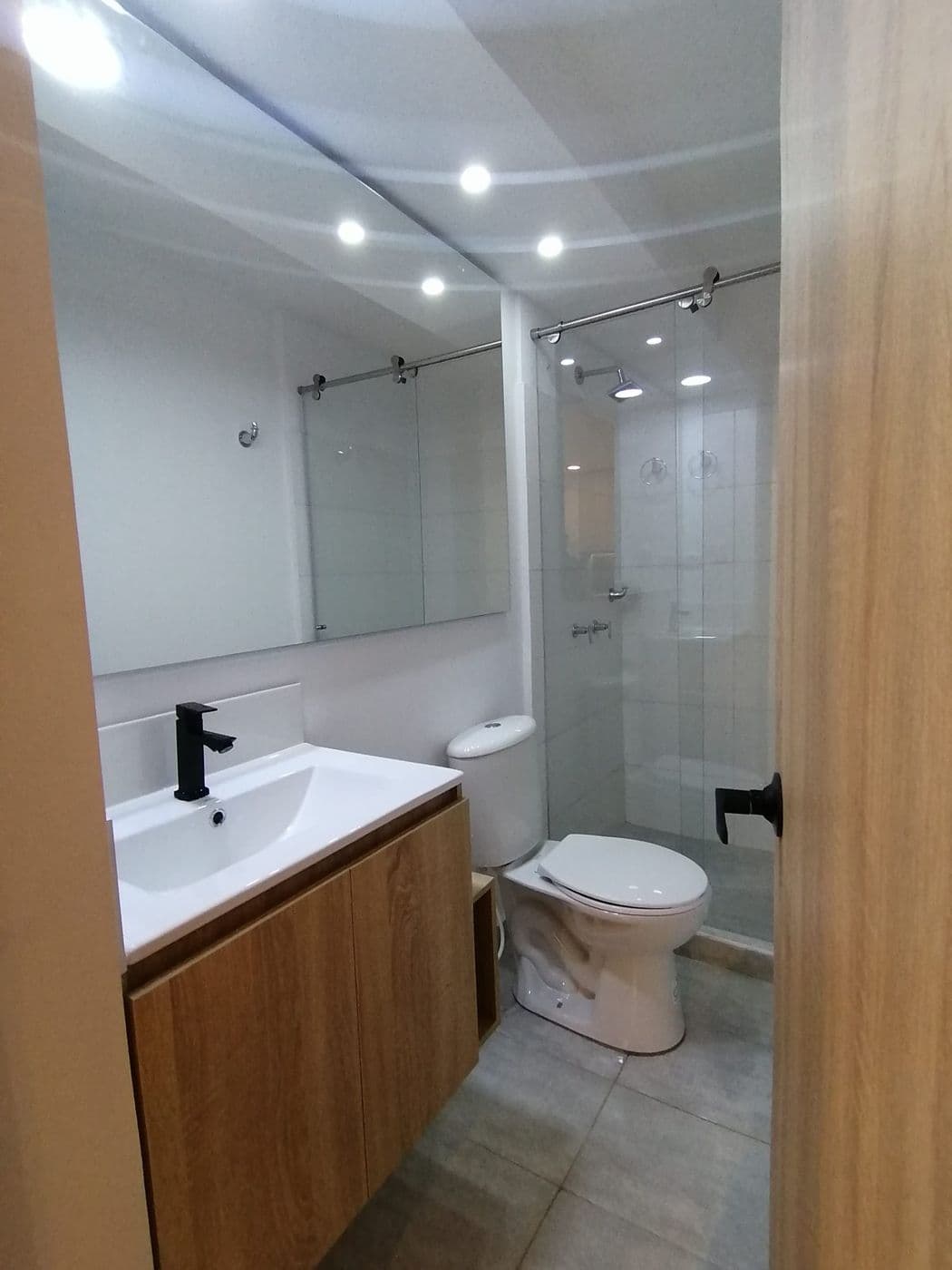 Baño con mueble flotante en madera clara, espejo amplio, ducha con puerta de vidrio corrediza y piso en porcelanato gris