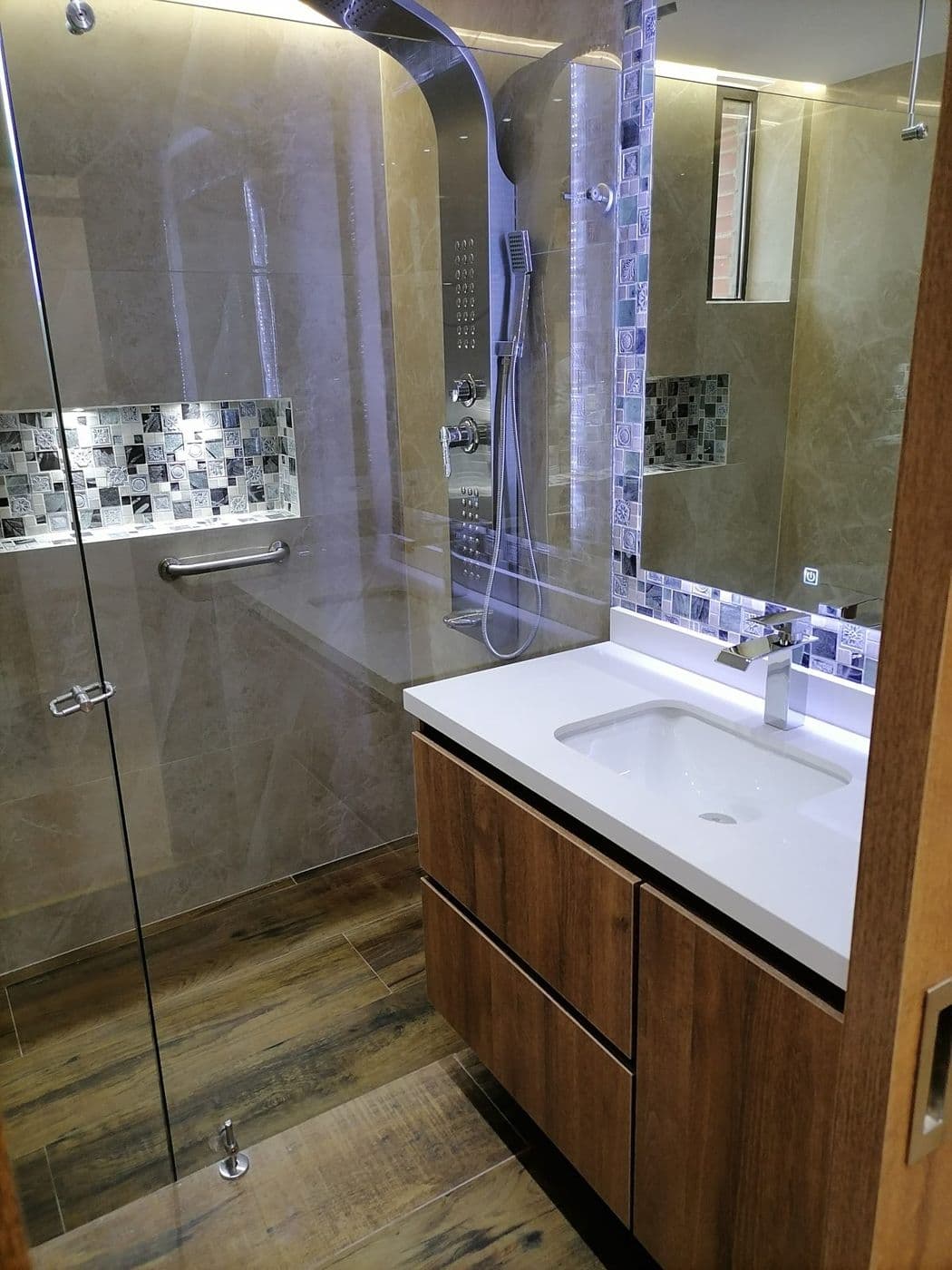 Ducha con enchape beige tipo mármol, franja de mosaico con luz LED y mueble de lavamanos en madera con mesón blanco