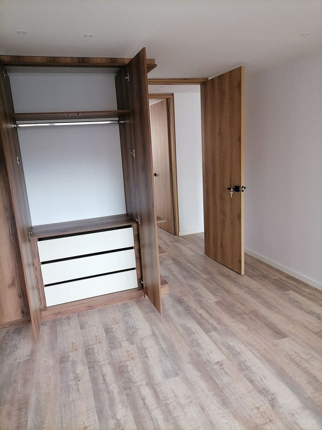 Closet en madera oscura abierto, con barra y cajones blancos, puerta a juego y piso laminado envejecido