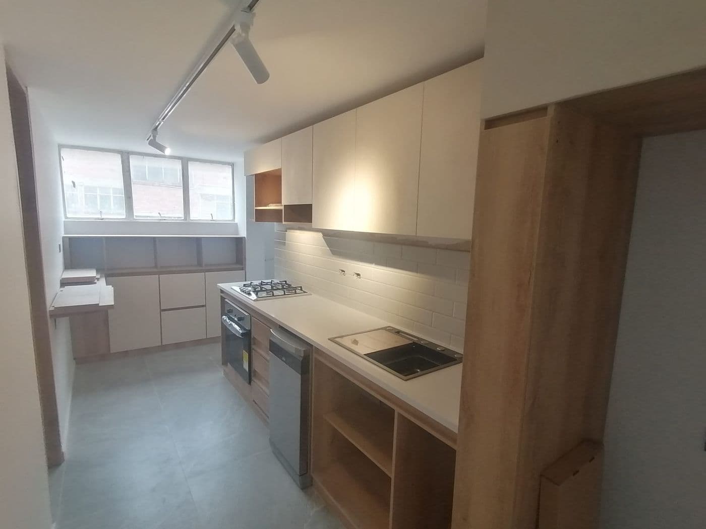 Cocina lineal con mesón blanco, salpicadero tipo ladrillo, muebles en madera y blanco, riel de luces y piso gris