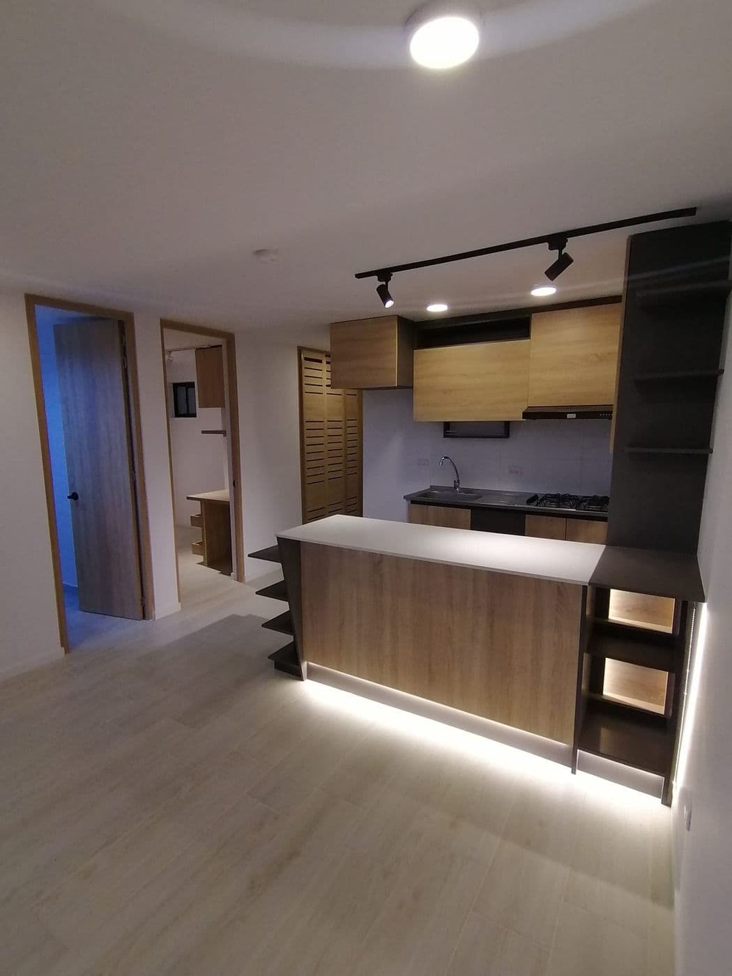Cocina abierta con barra de desayuno en madera clara, luz LED bajo la barra, repisas oscuras y piso tipo madera