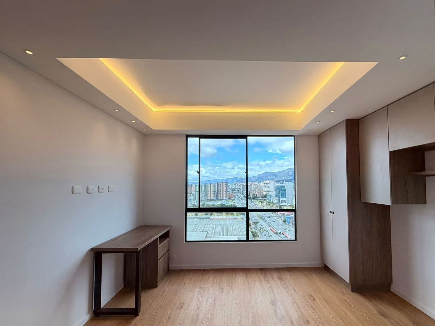 Estudio con escritorio en madera, cielo raso con luz indirecta cálida, ventanal y piso laminado