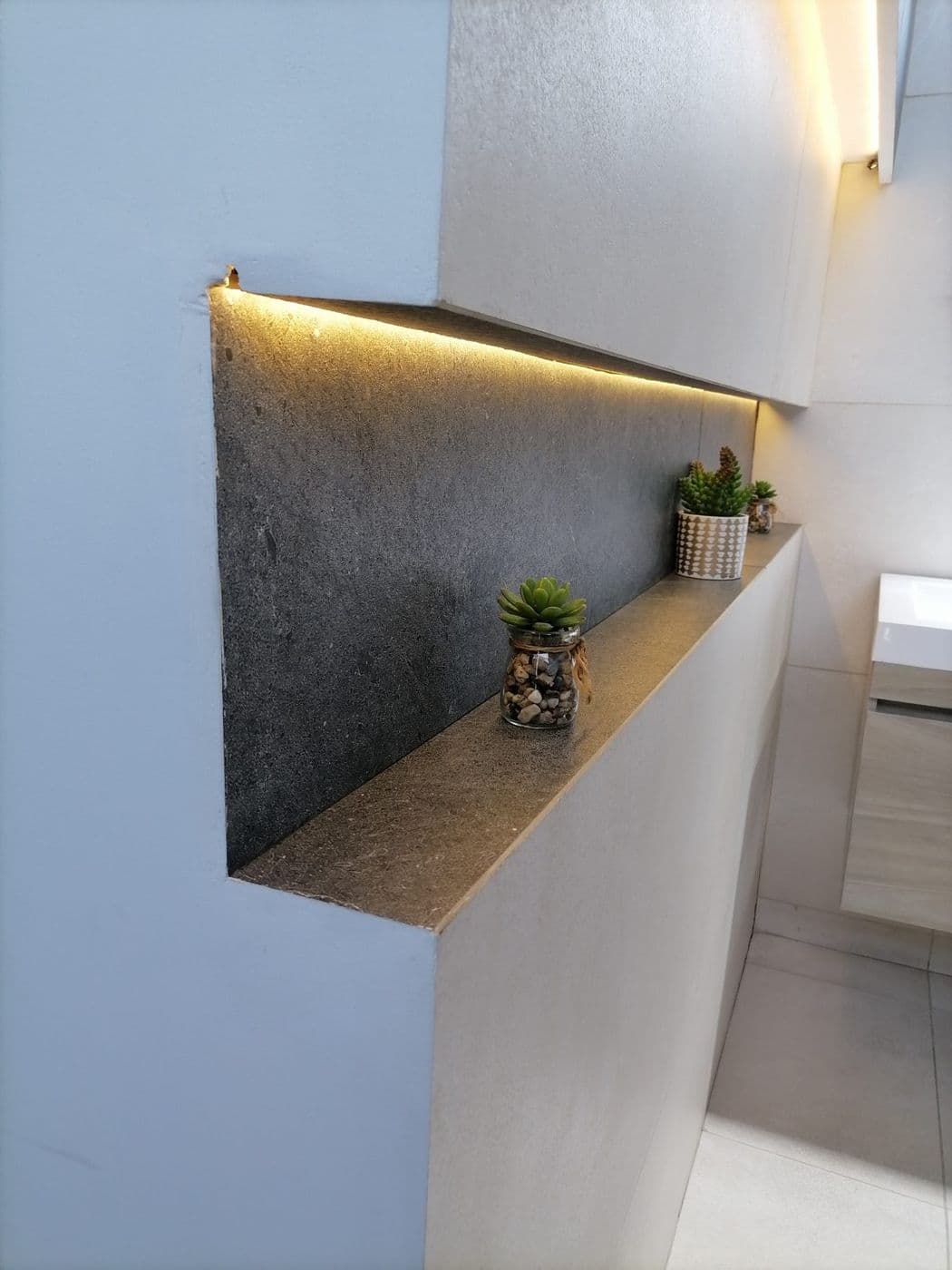 Nicho en enchape de piedra oscura con luz LED cálida y pequeñas plantas decorativas, junto a un mueble de lavamanos
